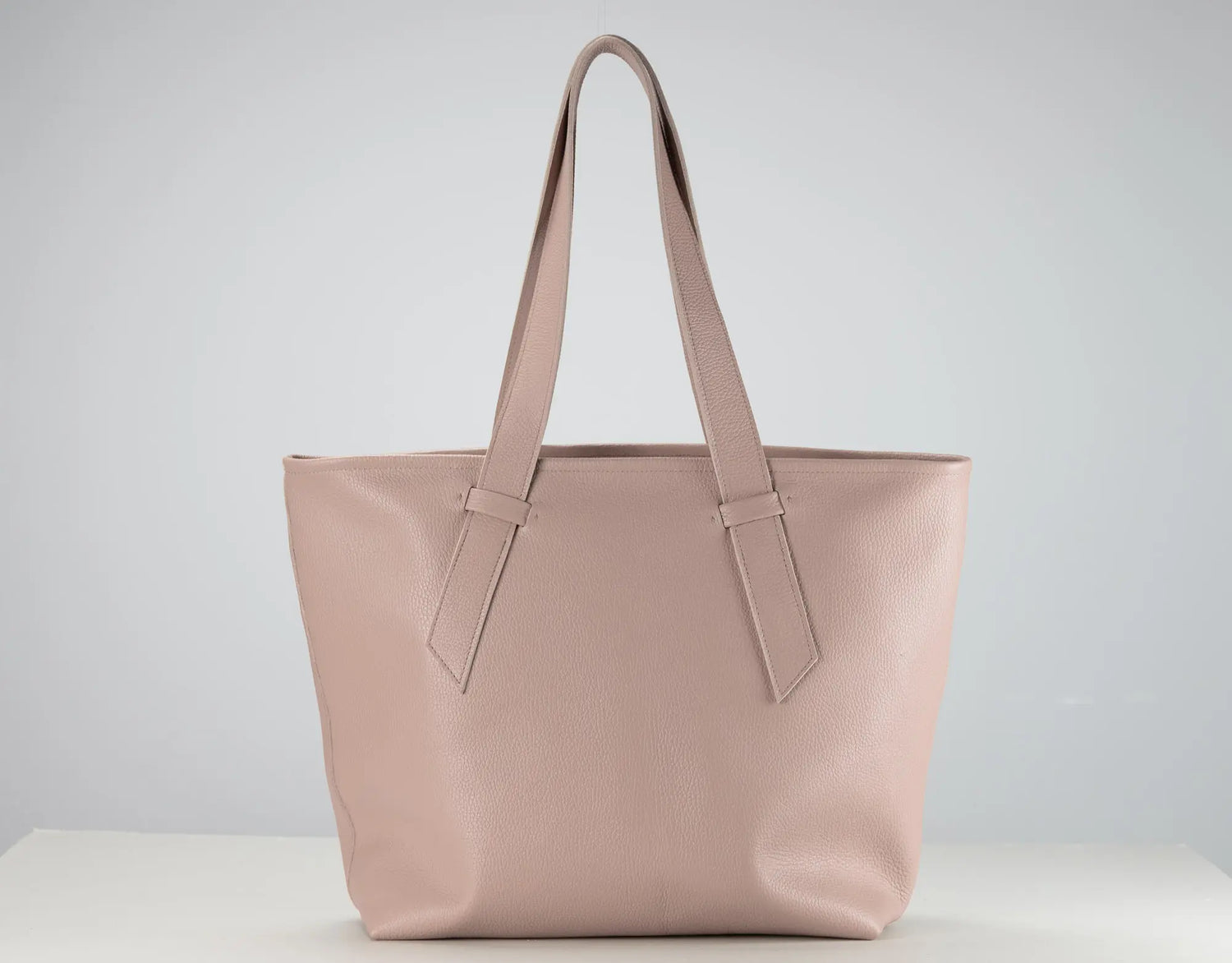 A light pink leather tote bag with dual shoulder straps, displayed on a white surface against a neutral light gray background.
