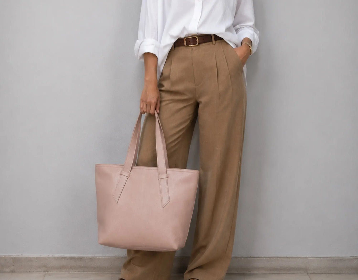 A person wearing a white button-up shirt, brown belt, and tan wide-leg trousers holds a large blush pink leather tote bag against a plain light gray wall.