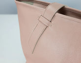 A close-up of a light blush pink pebbled leather bag, showing the texture of the leather and a matching strap detail.