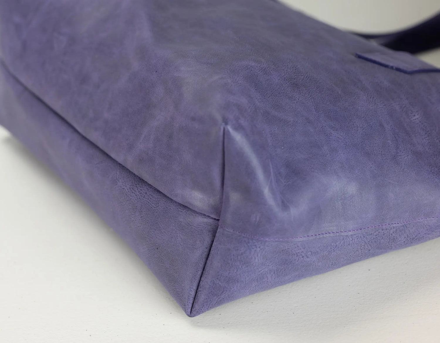 A close-up of a purple leather tote bag corner, showing its textured surface and stitched seam on a plain white background.