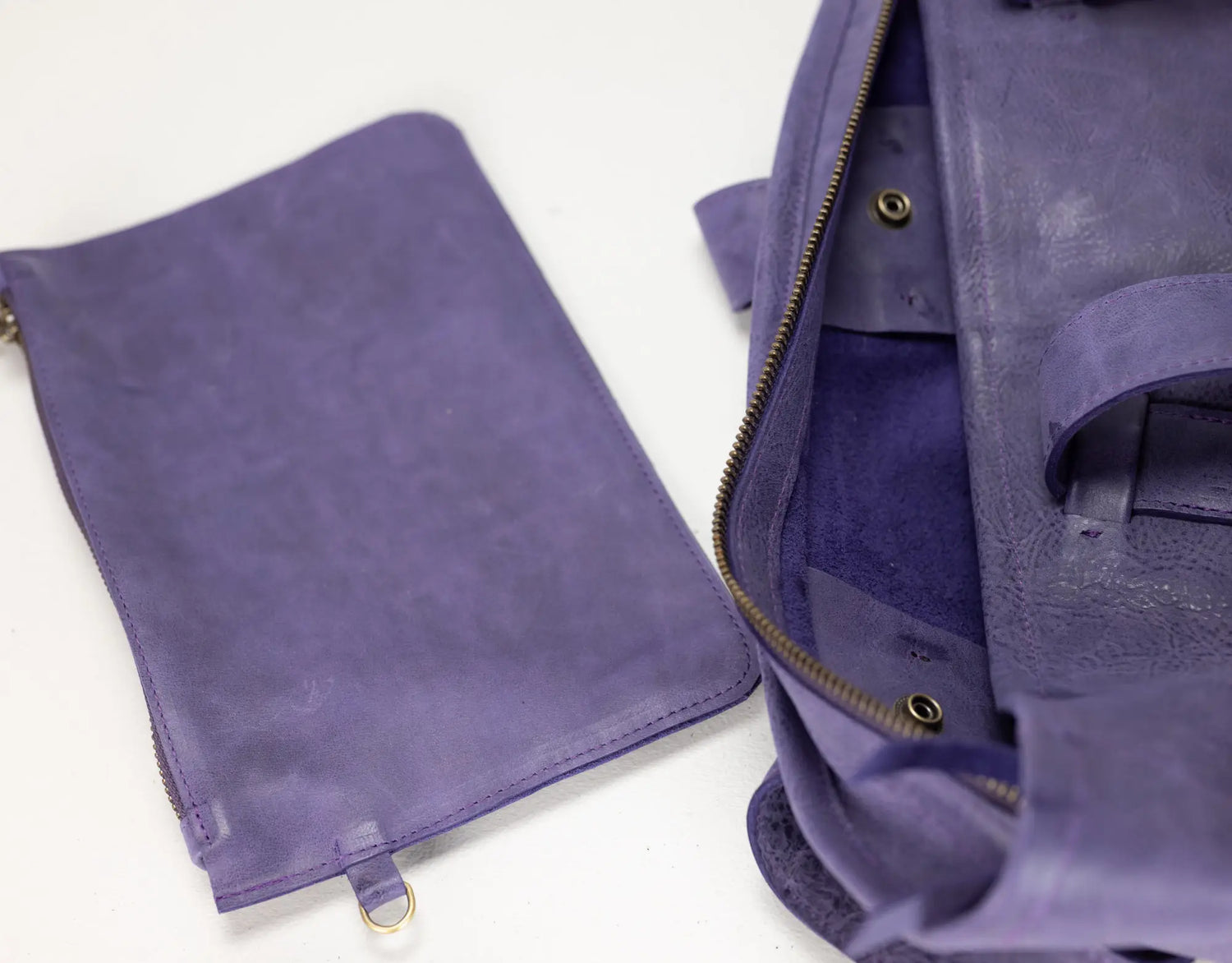 A purple leather zippered pouch shown next to an open matching purple bag, highlighting the interior and hardware on a plain white background.
