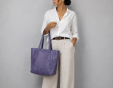 A woman wearing a white button-down shirt and cream trousers carries a large lavender-purple leather tote bag against a plain gray studio background.