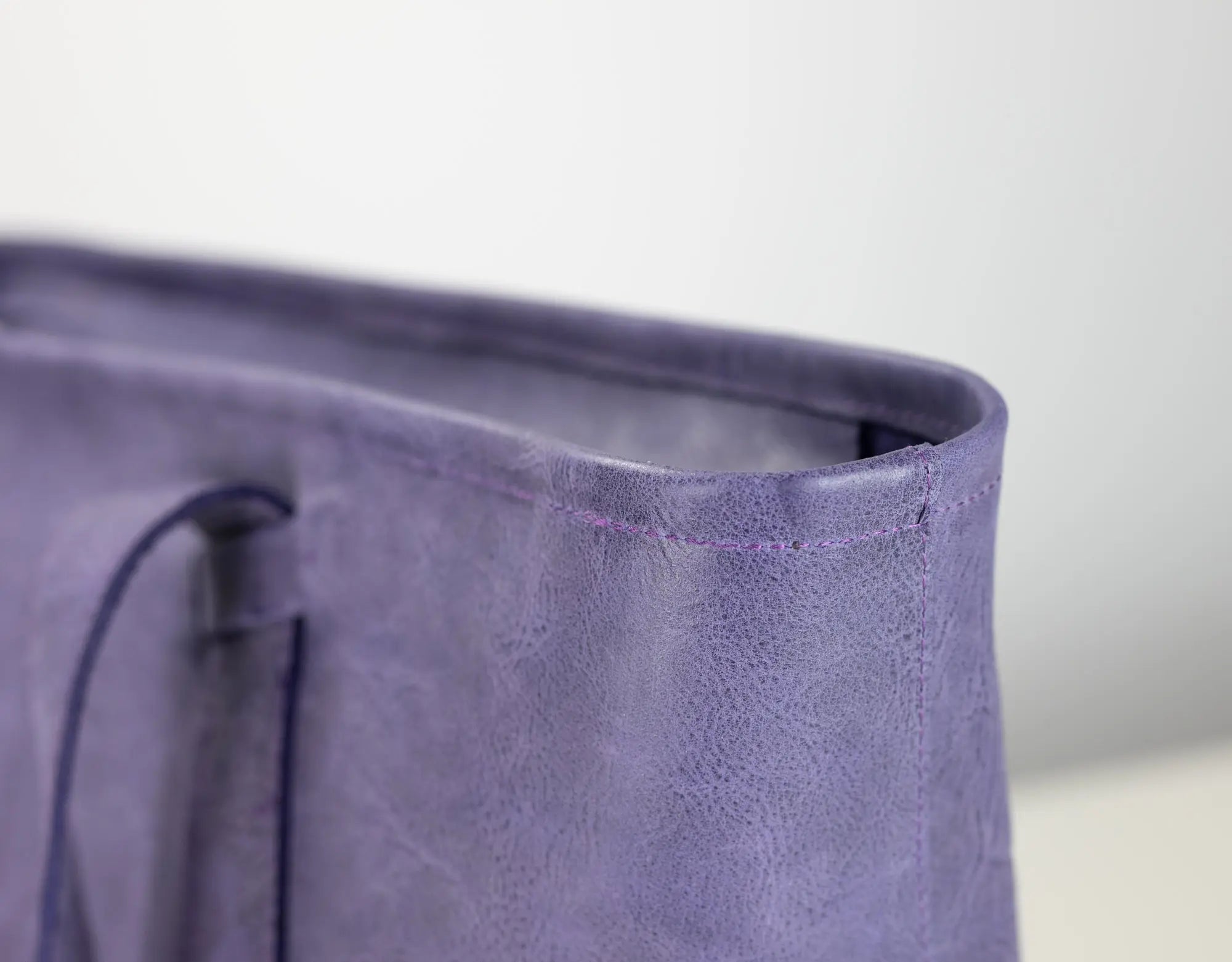 Close-up detail of a lavender purple textured leather tote bag corner, showing the stitched rim and part of the dark zipper pull.