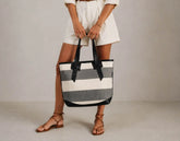 A person wearing white shorts and brown strappy sandals holds a large striped tote bag with black handles and black trim. The bag has horizontal stripes in off-white and gray, photographed against a plain light gray studio background.