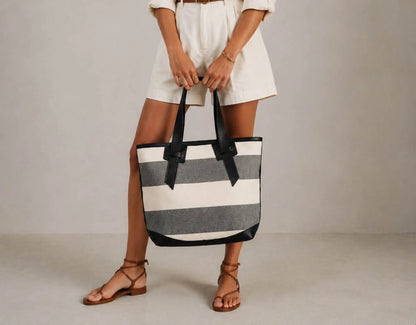 A person wearing white shorts and brown strappy sandals holds a large striped tote bag with black handles and black trim. The bag has horizontal stripes in off-white and gray, photographed against a plain light gray studio background.