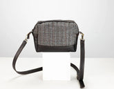 A small crossbody bag with a black leather base and trim, featuring a woven tweed pattern front panel in black, white, and gray. It has a long black leather shoulder strap and is displayed on a white block against a light gray background.