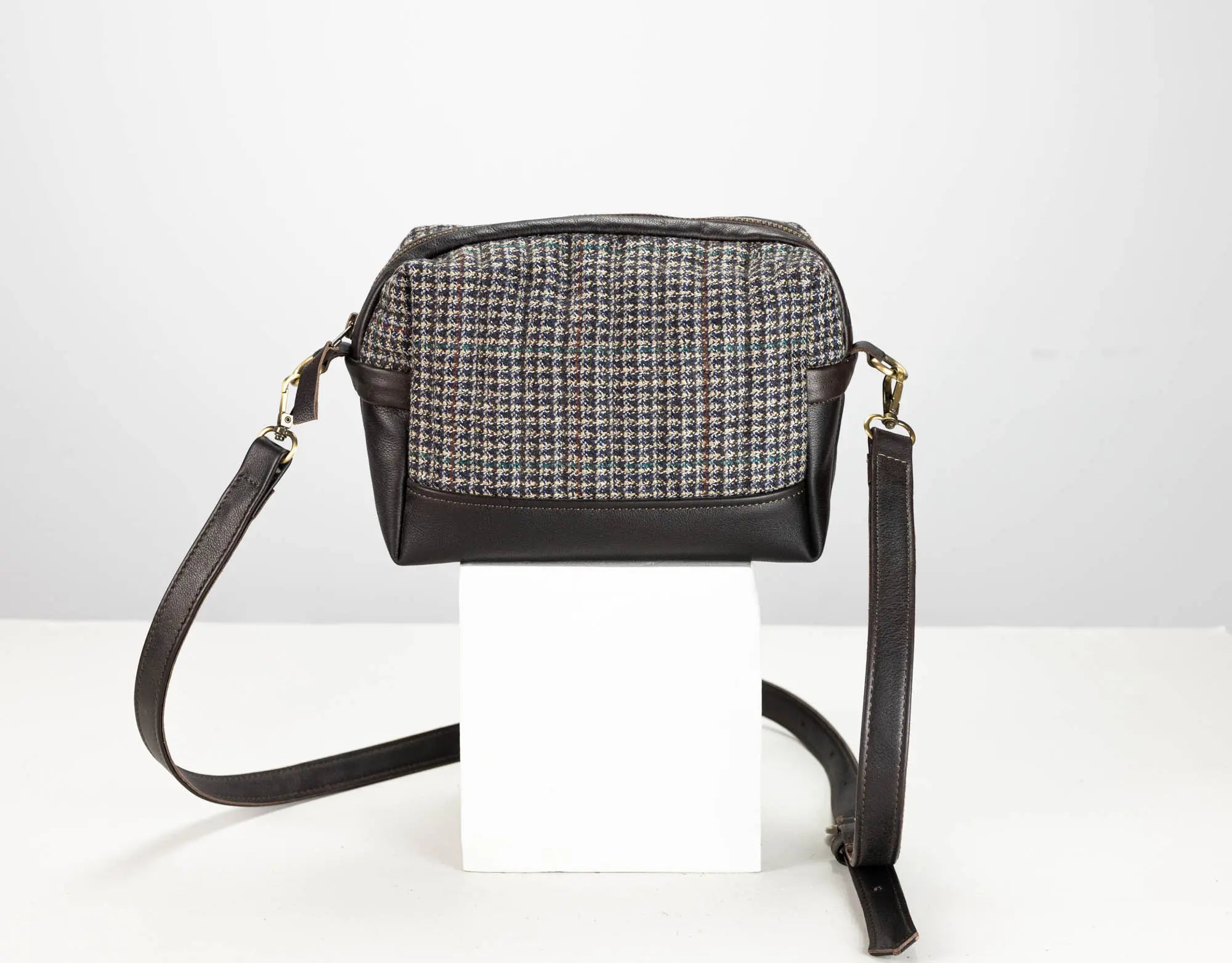 A small crossbody bag with a black leather base and trim, featuring a woven tweed pattern front panel in black, white, and gray. It has a long black leather shoulder strap and is displayed on a white block against a light gray background.