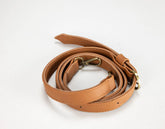 A tan faux-leather shoulder strap with brass-colored metal hardware, coiled on a plain white background.