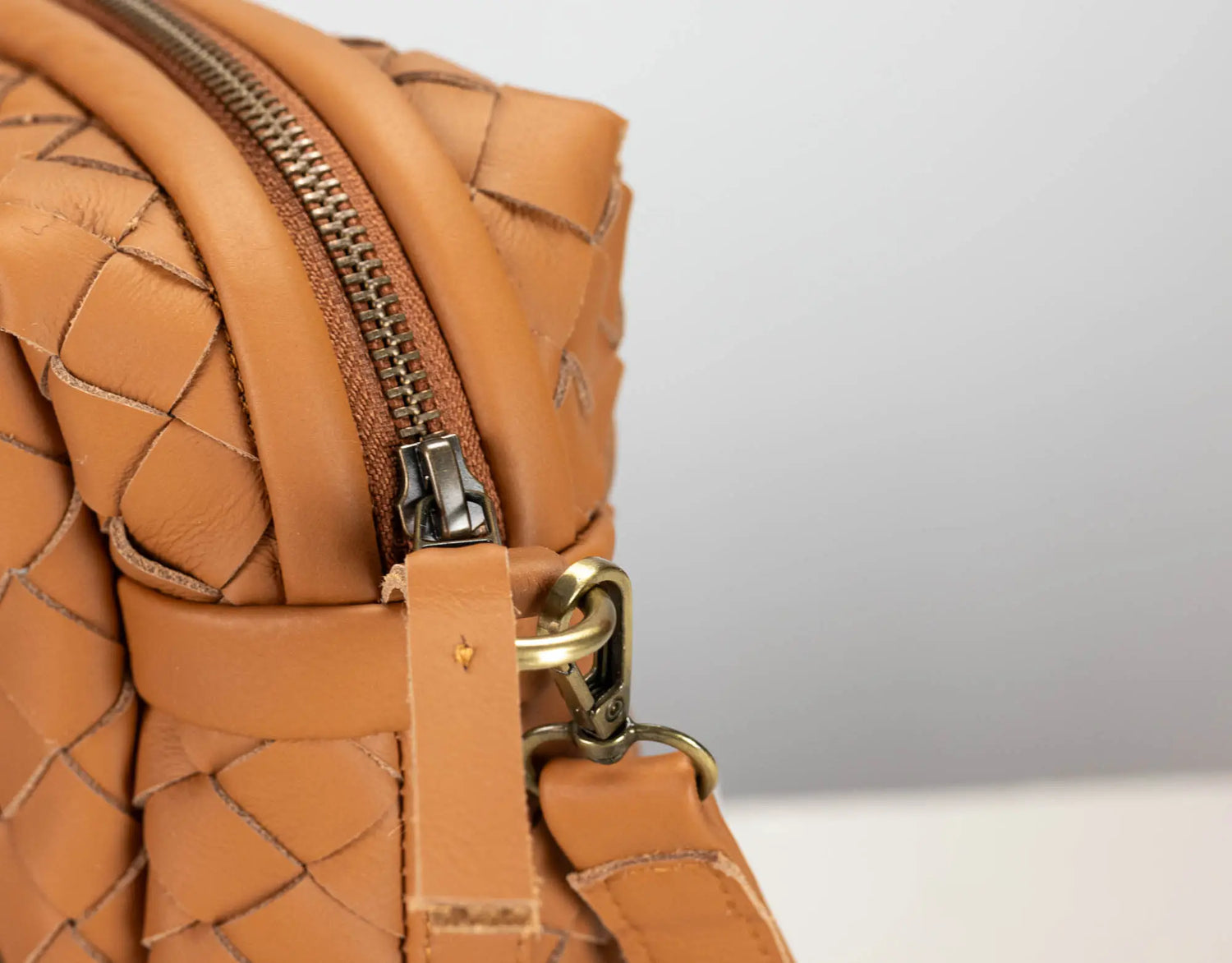 Close-up of a woven leather handbag in tan/caramel brown, showing the textured basketweave leather, a metal zipper, and brass-toned hardware against a light gray background.