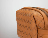 Close-up of a woven leather zippered pouch in a warm tan color, showing the distinctive zigzag stitching and metallic zipper against a light gray background.