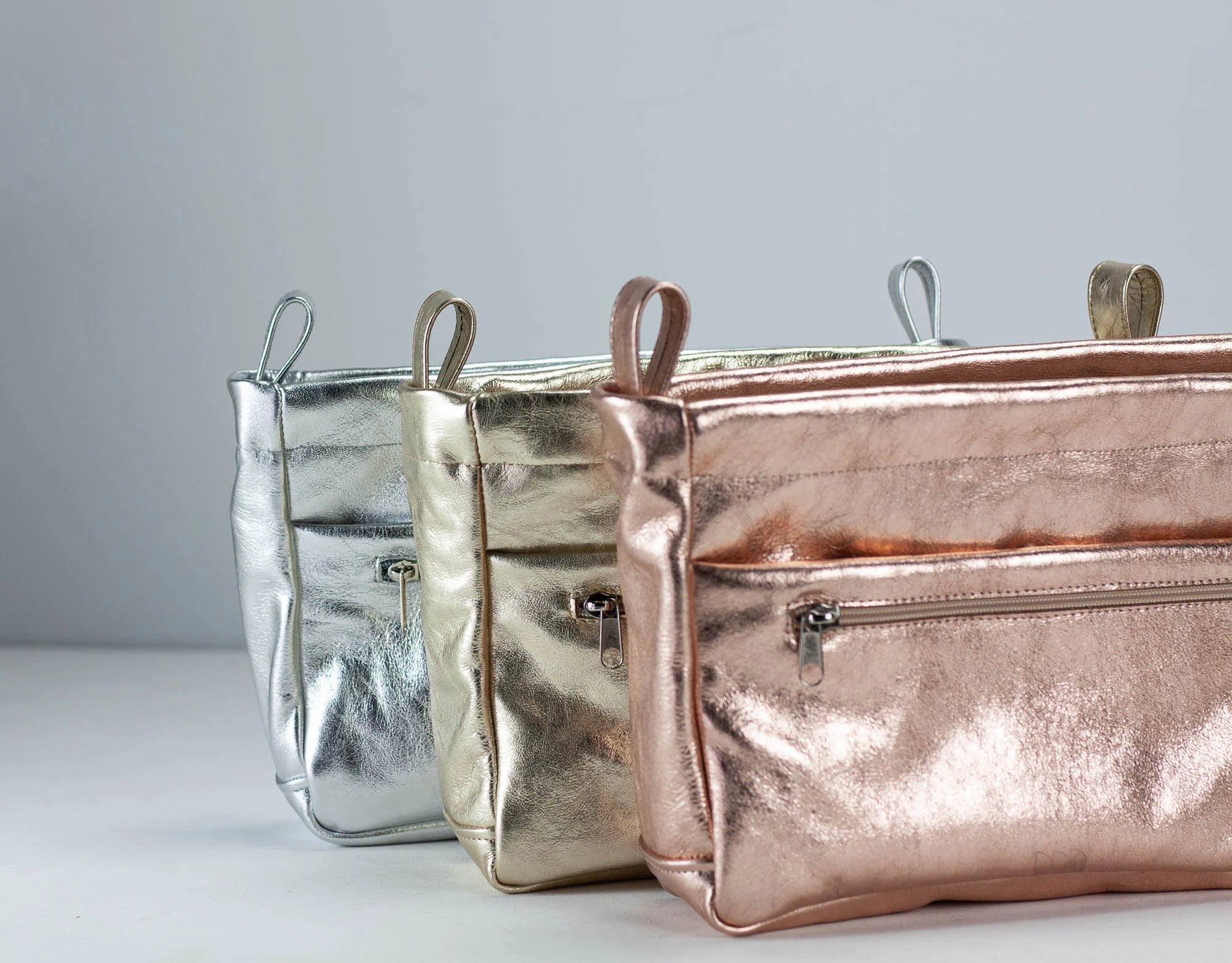 Three metallic handbags in silver, gold, and rose gold on a light gray background